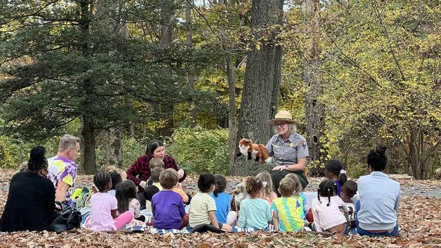 a group of young children sit on the forest floor listening to a park ranger with a fox hand puppet