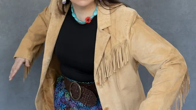Portrait of Danielle Soames, she wears a tan blazer with fringe along the arms and across the chest, a black shirt, and colorful skirt, she appears to be in the middle of a movement with an expression of apprehension