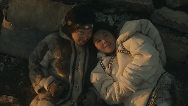 Film still from "Uiksaringitara (Wrong Husband)" of two people huddled close together against a rocky outcropping, one in a dark brown parka and gloves, the other in a white parka, both have traditional Inuit facial tattoos
