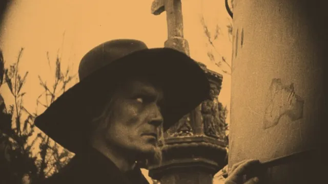 Sepia film still. Man with a craggy face and dark brimmed hat glowers amid gravestones.