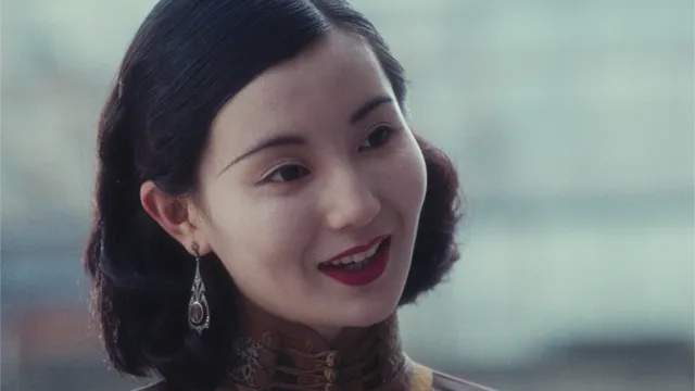 Close up of woman’s face mid-speech, head cocked with slight smile. She wears lipstick, ornate earrings, and a qipao.