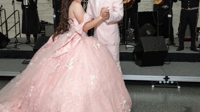 A quinceañera in a pink floral ball gown dances with her escort, who wears a pale pink suit, as a mariachi ensemble performs behind them.