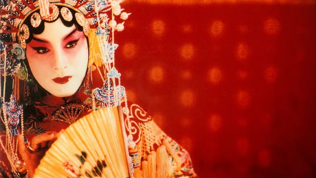A figure in feminine stage makeup and ornate headdress holds a folding fan and stares, a deep red background beyond.