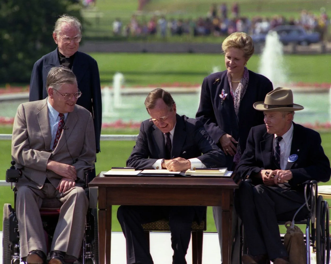 President George H. W. Bush signs the Americans with Disabilities Act on the White House South Lawn, seated between Evan Kemp and Justin Dart, with Rev. Harold Wilke and Swift Parrino standing behind.