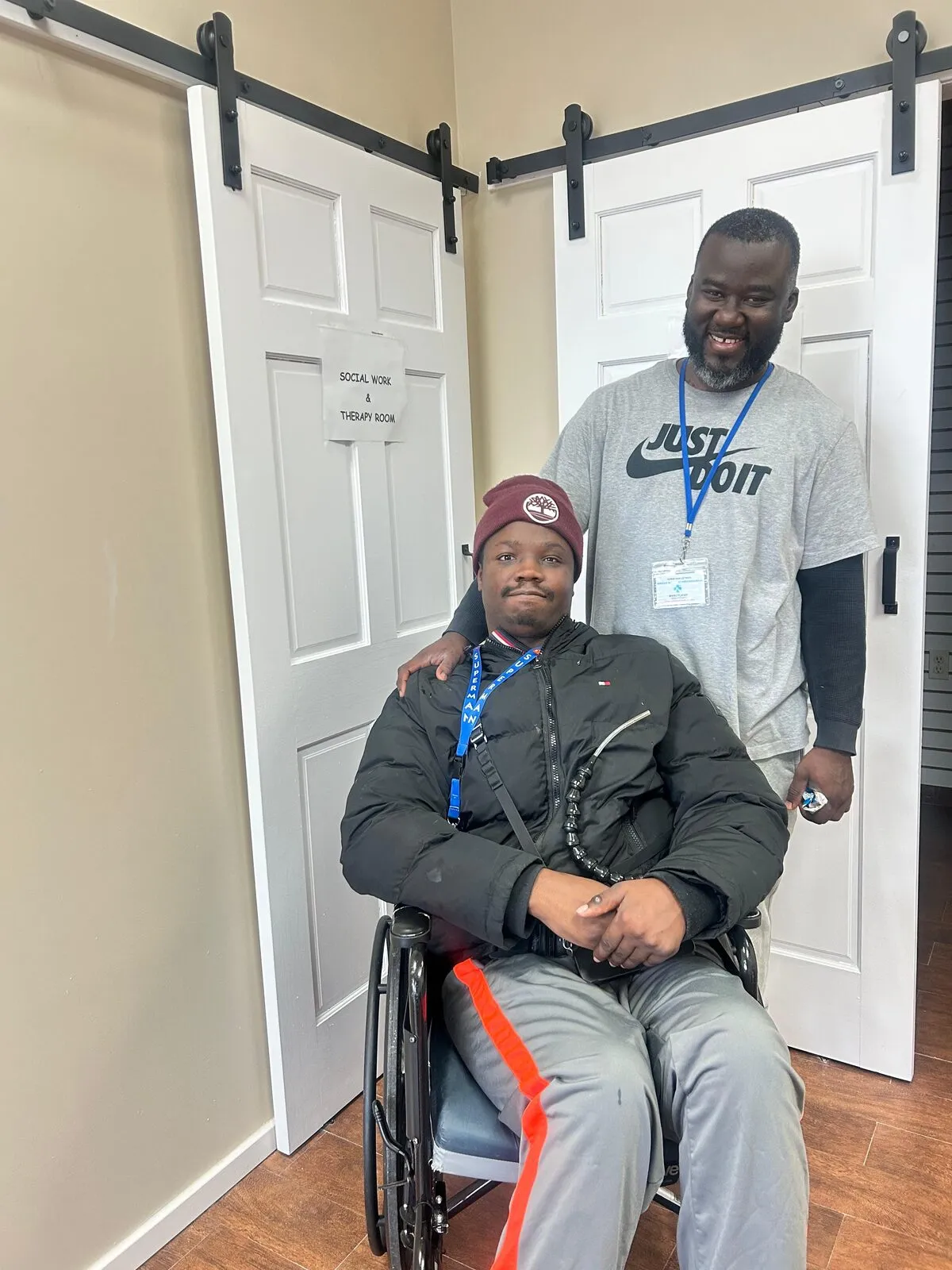 Jean-Christian stands beside Rasheed, seated in his wheelchair at home.