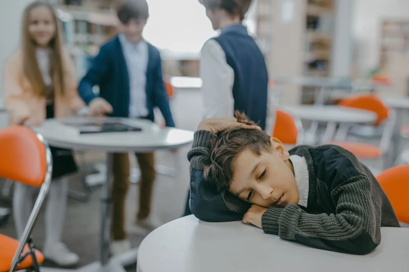 Un niño apoya la cabeza en una mesa del aula mientras otros estudiantes conversan detrás.