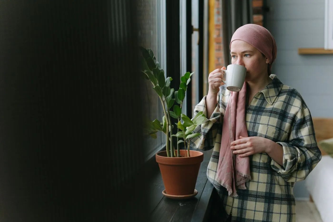 Una mujer con un turbante está junto a una ventana con una taza, mirando hacia afuera.
