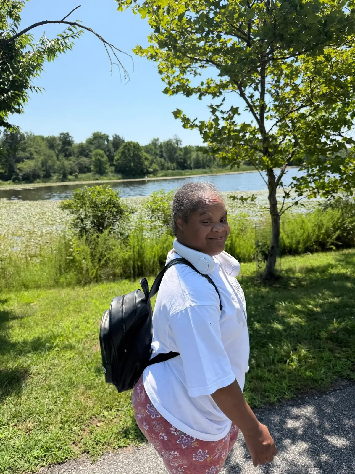 Ms. Rolanda walks along a park path by a lake, wearing a backpack.