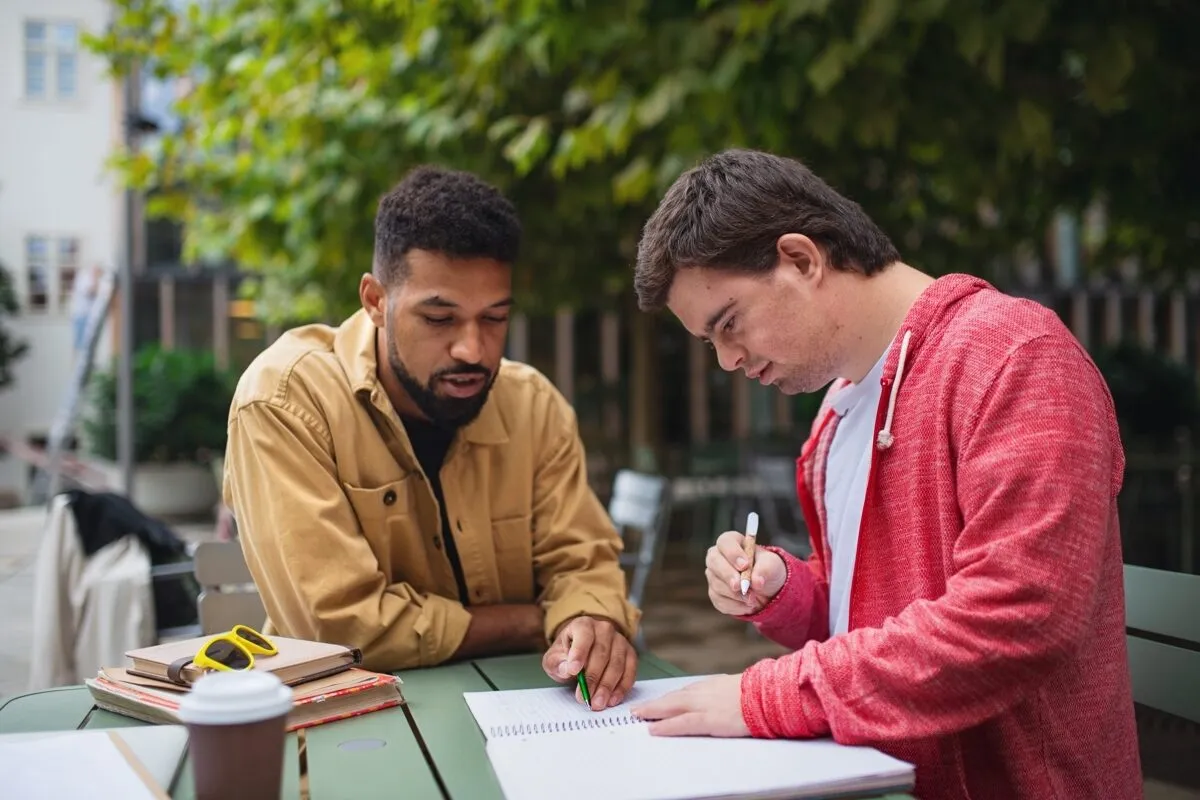 A support professional and a young man talk and work through notes together at an outdoor table.