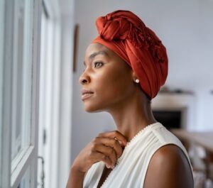 A woman in a red head wrap looks out of a window, one hand resting at her collarbone.