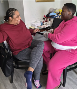 Una Profesional de Apoyo Directo de Mercyland, con uniforme rosa, se sienta frente a la mujer que apoya durante una visita a domicilio.