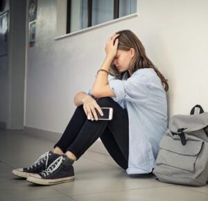 A young person sits alone on a hallway floor, head resting in one hand, a backpack beside them.