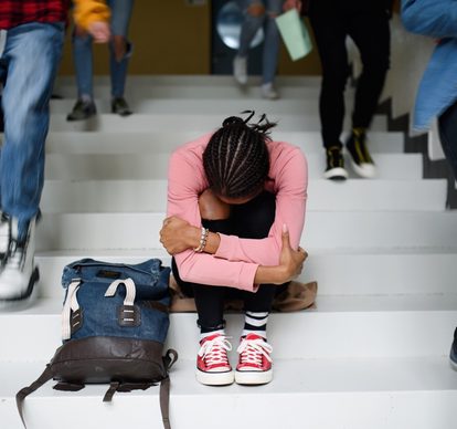 A student sits alone with their head down on a staircase, a backpack beside them, as other students walk past.