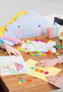 Un niño y una persona adulta trabajan juntos en una mesa cubierta de letras de espuma de colores y un dibujo, formando una palabra.
