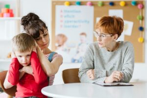 Una madre consuela a un niño que se tapa los oídos mientras una profesional toma notas al otro lado de la mesa.