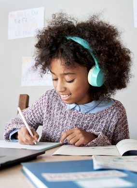 Una niña con audífonos que reducen el ruido sonríe mientras escribe en un escritorio rodeada de libros.